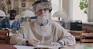 Portrait of stylish guy wearing eyeglasses sitting in library hall at desk studying looking at camera and smiling