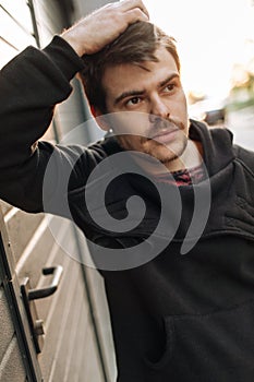 Portrait of stylish handsome young man with bristle standing outdoors and leaning on wall. Man wearing black hoody