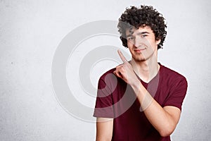 Portrait of stylish handsome young teenager isolated on white background. Dark haired curly student smiles and points aside.