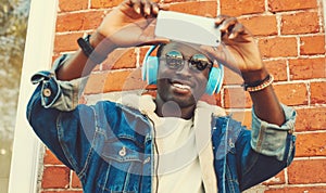 Portrait of stylish happy smiling african young man taking selfie with phone listening to music in headphones on city street