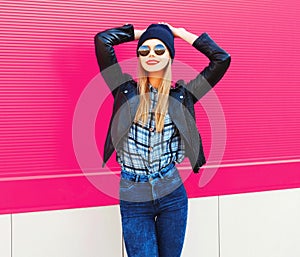 portrait stylish happy smiling woman in rock black style jacket, hat posing on city street over colorful pink wall