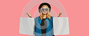 Portrait of stylish happy smiling woman with shopping bags wearing blue fur coat, black round hat and sunglasses posing on pink