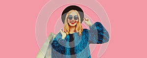 Portrait of stylish happy smiling woman with shopping bags wearing blue fur coat, black round hat and sunglasses posing on pink