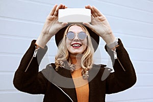 Portrait stylish happy smiling woman taking selfie picture by smartphone wearing black coat and hat on city street