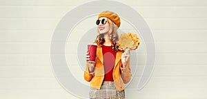 Portrait stylish happy smiling woman with yellow maple leaves, coffee cup in french beret posing over gray wall