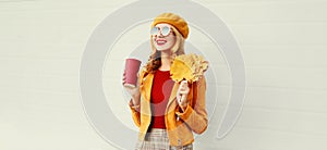 Portrait stylish happy smiling woman with yellow maple leaves, coffee cup in french beret posing over gray wall