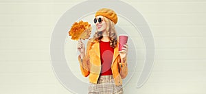 Portrait stylish happy smiling woman with yellow maple leaves, coffee cup in french beret posing over gray wall