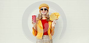 Portrait stylish happy smiling woman with yellow maple leaves, coffee cup in french beret posing over gray wall