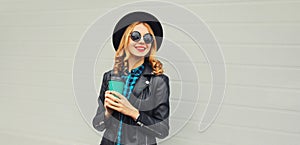 Portrait of stylish happy smiling young woman holds cup of coffee wearing round hat and rock style black leather jacket posing on