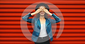 Portrait of stylish happy surprised smiling young woman covering her eyes wearing black round hat, blue leather jacket on red