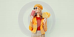 Portrait stylish happy woman drinking coffee with yellow maple leaves wearing french beret posing over gray wall