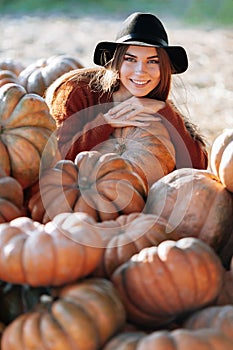 Portrait of stylish happy woman with smile posing among ripe orange pumpkins on farmers market in brown sweater and hat