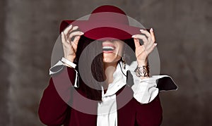 Portrait of stylish high-society lady in black jacket and white shirt pulling down hat brim hiding her eyes and laughs