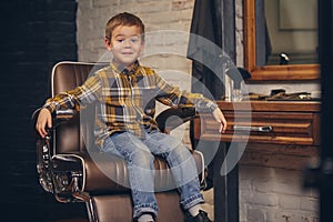 Portrait of a stylish little boy dressed in shirt and jeans in the barbershop, sitting in a chair against the barber`s