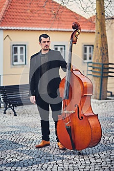 Portrait of a stylish man with a beard on an old street with a double bass. A solid musician with a large musical instrument in br