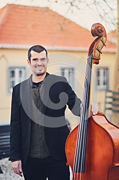 Portrait of a stylish man with a beard on an old street with a double bass. A solid musician with a large musical instrument in br