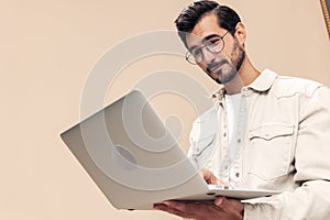 Portrait of a stylish man pensive look in glasses with a laptop in the hands of a freelancer, on a beige background in a