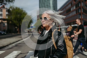 Portrait of stylish mature woman with gray hair on city street. Older woman in sunglasses crossing a road on pedestrian