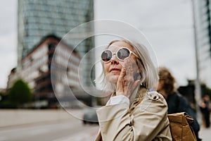 Portrait of stylish mature woman with gray hair on city street. Older woman in sunglasses waiting for public transport.