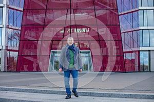 Portrait of stylish middle-aged man walking from modern building with glass facade in city outdoors, looking at camera.