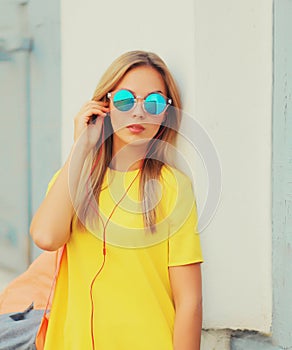 Portrait of stylish modern blonde young woman listening to music in headphones wearing sunglasses, backpack, yellow t-shirt in the