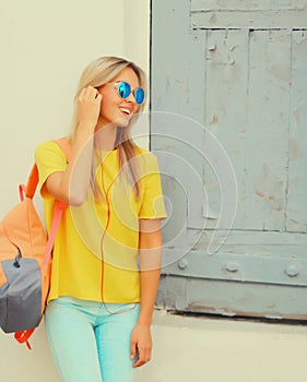 Portrait of stylish modern blonde young woman listening to music in headphones wearing sunglasses, backpack, yellow t-shirt in the