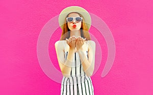 Portrait stylish pretty woman sending sweet air kiss in summer round straw hat, white striped jumpsuit on colorful pink wall