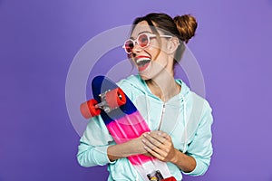 Portrait of stylish skater girl with two buns in sunglasses smiling and holding skateboard, isolated over violet background in st