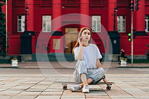 Portrait of a stylish skater woman in baggy jeans and white T-shirt sitting on a skateboard, looking at the camera. City street