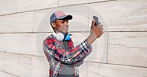 Portrait stylish smiling african man taking selfie picture by phone with headphones in baseball cap, plaid shirt on city street