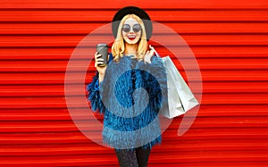 Portrait stylish smiling woman with coffee cup and shopping bags wearing blue faux fur coat, black round hat and sunglasses posing