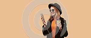 Portrait of stylish smiling woman looking at smartphone holding coffee cup wearing black coat, round hat isolated on brown