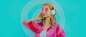 Portrait of stylish smiling young woman in wireless headphones listening to music wearing pink jacket, sunglasses over blue