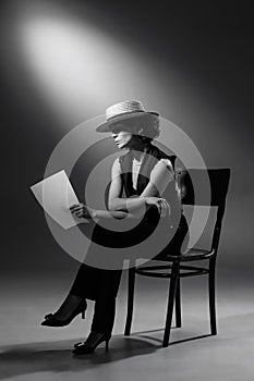 Portrait of stylish woman in classic black suit and straw hat, posing, sitting on chair and reading. Black and white