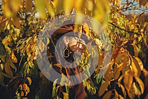 Portrait of stylish woman in hat and brown clothes posing among autumn leaves in warm sunny light. Fashionable young female