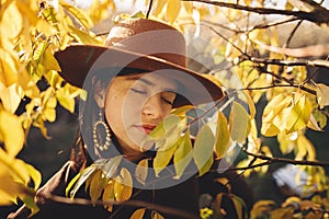 Portrait of stylish woman in hat and brown clothes posing among autumn leaves in warm sunny light. Fashionable young female