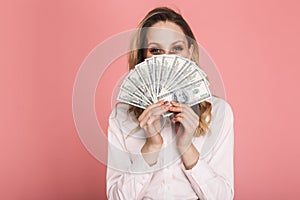 Portrait of stylish woman hiding while holding money fan isolated over pink background