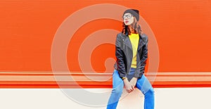 Portrait of stylish woman model posing wearing black rock style leather jacket, hat, eyeglasses on red background