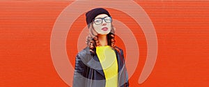 Portrait of stylish woman model posing wearing black rock style leather jacket, hat, eyeglasses on red background