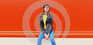 Portrait of stylish woman model posing wearing black rock style leather jacket, hat, eyeglasses on red background