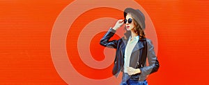 Portrait of stylish woman model posing wearing black rock style leather jacket, round hat on red background