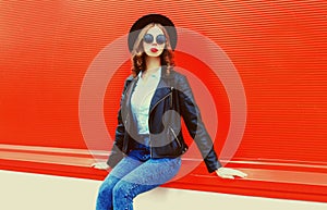 Portrait of stylish woman model posing wearing black rock style leather jacket, round hat on red background