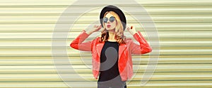 Portrait of stylish woman model posing wearing red rock style leather jacket, black round hat on gray background