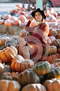Portrait of stylish woman posing among ripe orange pumpkins on farmers market in brown sweater, dress and hat. Cozy autumn vibes