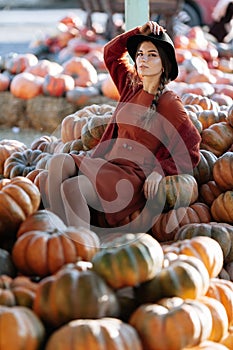 Portrait of stylish woman posing among ripe orange pumpkins on farmers market in brown sweater, dress and hat. Cozy autumn vibes