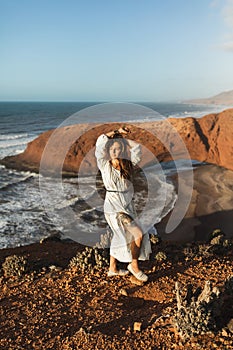 Portrait of stylish woman in white dress on Legzira beach in Morocco. Tattoo leg