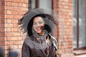 Portrait of stylish young African American woman smiling happily. Brunette with curly hair in brown leather jacket