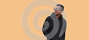 Portrait of stylish young african man looking away wearing black hoodie, sunglasses isolated on brown background, blank copy space