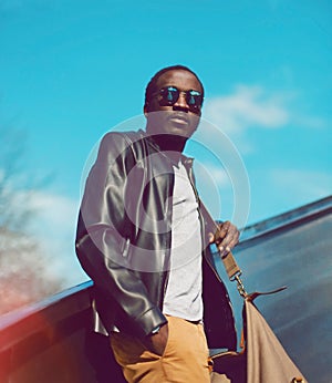 Portrait of stylish young african man model posing wearing black rock leather jacket with bag on city street background