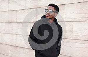 Portrait stylish young african man walking and looking away wearing a black hoodie, sunglasses on a city street over gray wall